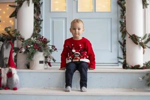 glücklich wenig Junge im Weihnachten. ein Kind im ein Weihnachten Sweatshirt sitzt auf das Veranda von ein dekoriert Zimmer. foto