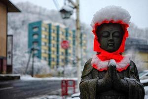 uralt Stein Buddha Bild mit rot Kapuze im fallen Schnee im noboribetsu Onsen wo ist berühmt Tourist Attraktion im Hokkaido, Japan. foto