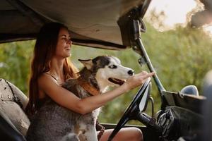 ein schön jung Frau sitzt hinter das Rad von ihr Auto zusammen mit ein heiser Rasse Hund und lächelt fröhlich genießt das Reise foto
