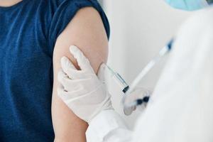 Frau Arzt im schützend Handschuhe Injektionen im das Schulter von ein Mann im ein Blau T-Shirt Impfung foto