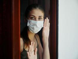 Frau mit traurig Gesichts- Ausdruck im medizinisch Maske hält Hände beim Sperrung Fenster foto