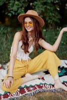 Hippie Frau lächelnd im Öko Kleidung Gelb Hose, Weiß stricken Spitze, Hut und Gelb Brille Sitzung auf Plaid im Park Aufpassen Sonnenuntergang, Lebensstil Camping Ausflug foto