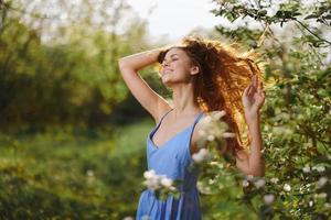 Frau mit ein schön Lächeln mit Zähne und lange Haar fliegend Haar im das Frühling Sonnenuntergang im Natur im das Park in der Nähe von das blühen Bäume Glück, natürlich Schönheit und Haar Gesundheit foto