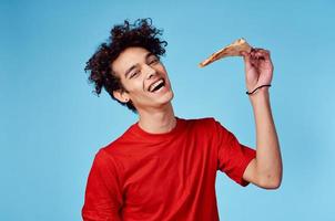 Mann mit ein Stück von Pizza im seine Hand auf ein Blau Hintergrund Teenager im ein rot T-Shirt lockig Haar foto