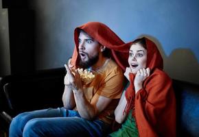 ein Mann mit ein Teller von Popcorn und ein emotional Frau unter ein rot Decke auf das Couch foto