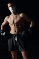 sportlich Mann im medizinisch Maske und im Boxen Handschuhe auf schwarz Hintergrund kurze Hose foto