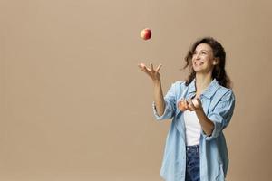 glücklich lächelnd konzentriert lockig schön weiblich im Jeans beiläufig Hemd Jonglieren mit Apfel posieren isoliert auf Über Beige Pastell- Hintergrund. gesund Lebensmittel. natürlich umweltfreundlich Produkte Konzept. Kopieren Raum foto