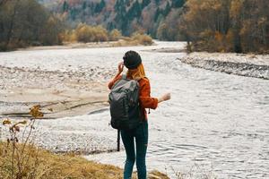 ein Reisender im ein Sweatshirt Hut mit ein Rucksack Gesten mit ihr Hände und entspannt sich auf das Fluss Bank im das Berge foto