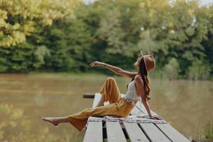 ein Hippie Frau sitzt auf ein Brücke auf das Bank von ein Fluss und genießt das schön Landschaft um ihr, glücklich im das Sonnenuntergang Herbst Sonne. ökologisch Lebensstil foto