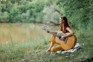 Hippie Frau spielen Gitarre lächelnd und Singen Lieder im Natur Sitzung auf ein Plaid durch das See im das Abend im das Strahlen von das Rahmen Sonne. ein Lebensstil im Harmonie mit das Körper und Natur foto