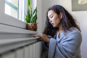 asiatisch Frau Einfrieren beim heim, Sitzung durch das kalt Kühler. Frau mit Zuhause Heizung Problem Gefühl kalt foto