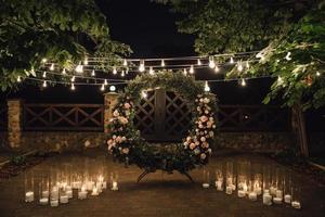 schön Photozone mit groß Kranz dekoriert mit Grün und Rosen im Kernstück, Kerzen auf das Seiten, und Girlande gehängt zwischen Bäume foto