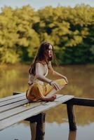 ein jung glücklich Frau mit ein Hippie Lächeln sitzt auf das See Ufer auf ein Brücke und meditiert im ein Lotus Pose tragen Öko Kleidung gemacht von natürlich Materialien im Harmonie mit Natur im das fallen foto