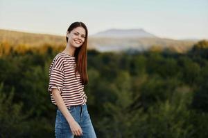 ein jung Frau lacht und sieht aus beim das Kamera im einfach Kleider gegen das Hintergrund von ein schön Landschaft von Berge und Bäume im Herbst. Lebensstil auf das Bewegung foto