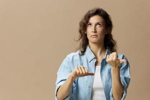 verwirrt lockig schön weiblich im Jeans beiläufig Hemd können nicht machen Entscheidung Show Daumen oben Nieder sieht aus oben posieren isoliert auf Über Beige Pastell- Hintergrund. Menschen Lebensstil Emotionen Konzept. Kopieren Raum foto