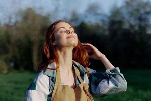 Porträt von ein schön jung Frau mit rot Haar im Natur im ein Park im das Sommer, suchen beim das Sonnenuntergang gegen ein Hintergrund von Grün Gras und Bäume foto