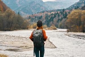 glücklich Frau Wanderer mit ein Rucksack auf das Fluss Bank aussehen in das Berge und Herbst Wald Natur foto