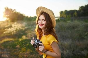 Frau Fotograf mit ein Kamera im ihr Hände Lächeln rot Lippen Hut Gelb T-Shirt Sommer- Natur foto