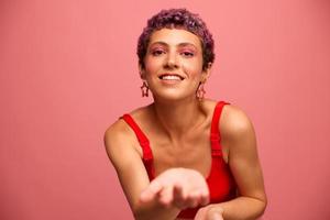 Mode Porträt von ein Frau mit ein kurz Haarschnitt von lila Farbe und ein Lächeln mit Zähne im ein rot oben auf ein Rosa Hintergrund Tanzen glücklich foto