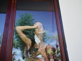 Frau halten ihr Kopf im das Fenster tragen ein medizinisch Maske Quarantäne foto