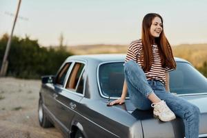 ein jung Frau sitzt auf das Kofferraum von ein Auto und ruht nach ein schwierig Straße und bewundert Natur mit ein schön Sicht. anhalten ist ebenfalls Teil von das Reise foto