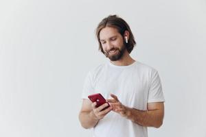ein Mann mit ein Bart im Weiß T-Shirt und Jeans sieht aus beim seine Telefon umdrehen durch ein online Sozial Medien Futter mit Kopfhörer im seine Ohren Hören zu ein Stimme Botschaft auf ein Weiß Hintergrund und lächelnd foto
