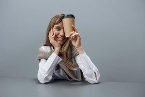 heiter jung blond Schüler Dame Freiberufler im warm Sweatshirt mit wegbringen Kaffee Lächeln beim Kamera Sitzung beim Arbeitsplatz im grau modern Zuhause Büro. Kaffee brechen Liebhaber Konzept. Kopieren Raum. cool Angebot foto