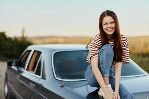 ein jung Frau sitzt auf das Kofferraum von ein Auto, lacht und ruht nach ein schwierig Straße und bewundert Natur mit ein schön Sicht. anhalten ist ebenfalls Teil von das Reise foto
