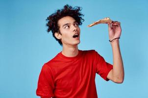 Mann mit ein Stück von Pizza im seine Hand auf ein Blau Hintergrund Teenager im ein rot T-Shirt lockig Haar foto