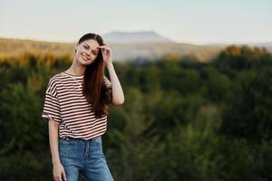 ein jung Frau lacht und sieht aus beim das Kamera im einfach Kleider gegen das Hintergrund von ein schön Landschaft von Berge und Bäume im Herbst. Lebensstil auf das Bewegung foto