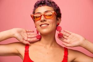 jung sportlich Frau mit ein kurz Haarschnitt und lila Haar im ein rot oben und Rosa Yoga Gamaschen im Sonnenbrille mit ein sportlich Zahl lächelt und Tänze auf ein Rosa Hintergrund foto