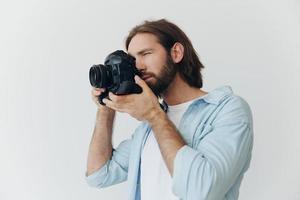 ein männlich Hipster Fotograf im ein Studio gegen ein Weiß Hintergrund sieht aus durch das Kamera Sucher und schießt Schüsse mit natürlich Licht von das Fenster. Lebensstil Arbeit wie ein freiberuflich Fotograf foto