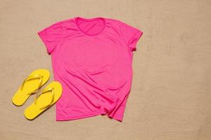 Sand Strand Textur Hintergrund. Attrappe, Lehrmodell, Simulation Rosa Sommer- T-Shirt Kopieren Raum. leer Vorlage Frau Hemd oben Sicht. Sommer Zubehör Flip-Flops. flach liegen Nahansicht T-Shirt auf Strand. Strandzeit foto