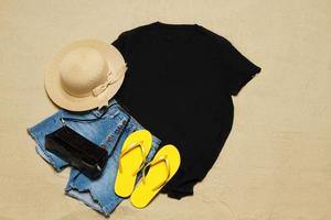 Sand Strand Textur Hintergrund. Attrappe, Lehrmodell, Simulation schwarz Sommer- T-Shirt Kopieren Raum. leer Vorlage Frau Hemd oben Sicht. Sommer Zubehör Hut, Jeans, Flip-Flops. flach liegen Nahansicht T-Shirt Strand. Strandzeit foto