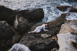 barfuß Frau im ein Hochzeit Kleid Lügen auf ein Stein im ein Weiß Kleid Lügen auf ihr zurück auf ein felsig Strand foto