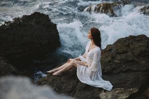 ein Frau im ein Weiß Kleid barfuß sitzt auf ein Stein auf ein Cliff oben Aussicht unverändert foto