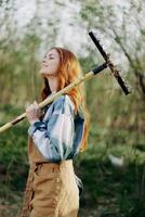 ein Frau Farmer im Arbeit Kleider und ein Schürze funktioniert draußen im Natur und hält ein Rechen zu versammeln Gras foto