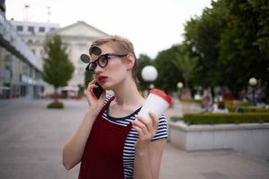 Frau mit Brille auf das Straße reden auf das Telefon im ein Glas mit ein trinken foto