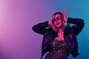 sexy schön blond Dame im Leder Jacke prickelnd Kleid modisch Sonnenbrille berühren Kopf beide Hände posieren isoliert im Blau Rosa Farbe Licht Studio Hintergrund. Neon- Party Cyberpunk Konzept. Kopieren Raum foto