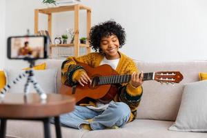Blogger Gitarrist. glücklich afrikanisch amerikanisch Mädchen Blogger spielen Gitarre Singen Lied Aufzeichnung vlog. Sozial Medien Influencer Frau Streaming Aufzeichnung beim Zuhause Studio. Musik- Inhalt Schöpfer Übertragung. foto