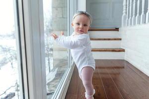 Kleines krabbelndes Mädchen, ein Jahr alt, sitzt lächelnd und lachend auf dem Boden im hellen Wohnzimmer in der Nähe des Fensters foto