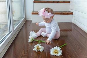 wenig Baby Mädchen tragen Frühling Kranz Standort auf Fußboden im hell Licht Leben Zimmer in der Nähe von Fenster und spielen mit Gerbera Blumen foto