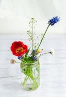 kleiner Strauß der Frühlingsfeldblumen in einem Glasglas auf weißem rustikalem Holzhintergrund foto