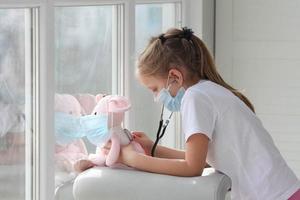 wenig Mädchen im medizinisch Maske halten Stethoskop Hören zu krank Spielzeug, Plüsch Ratte, geduldig, Clever wenig Kind spielen Krankenhaus Spiel mögen Arzt vortäuschend, Tierarzt. foto