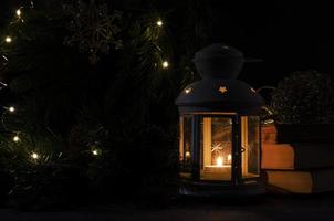 Weiß Laterne mit ein Verbrennung Kerze. Bücher, Weihnachten Baum Ast und Beleuchtung auf Hintergrund. Nacht dunkel Bild. Weihnachten Geheimnis Stimmung. foto