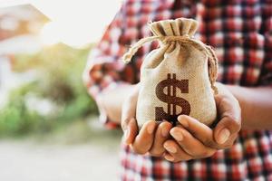 Konzept Finanzen Buchhaltung. Hand halten Geld Tasche mit Sonnenuntergang und Haus Hintergrund foto