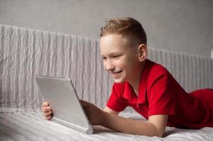 ein süß Junge im ein rot T-Shirt ist Lügen und spielen auf ein Tablette auf das Couch und Lachen foto