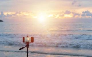 Jemand, der am Patong Beach, Phuket, Thailand mit einem Handy auf einem Stativ reist und auf den Sonnenuntergang wartet, um ein gutes Foto zu machen