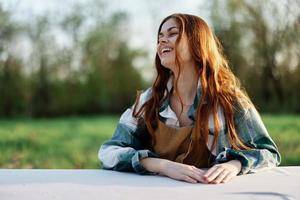 ein schön Frau lacht mit ein zahnig Lächeln und genießt faulenzen im ein Grün Park auf ein Sommer- Abend im das Rahmen Sonnenlicht. das Konzept von ein gesund Lebensstil und das bati von Selbstpflege foto