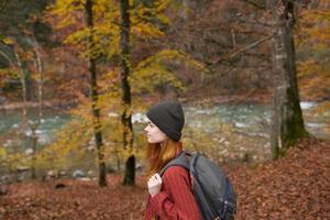 Seite Aussicht von glücklich Frau im Park im Herbst in der Nähe von Fluss und Rucksack auf ihr zurück foto